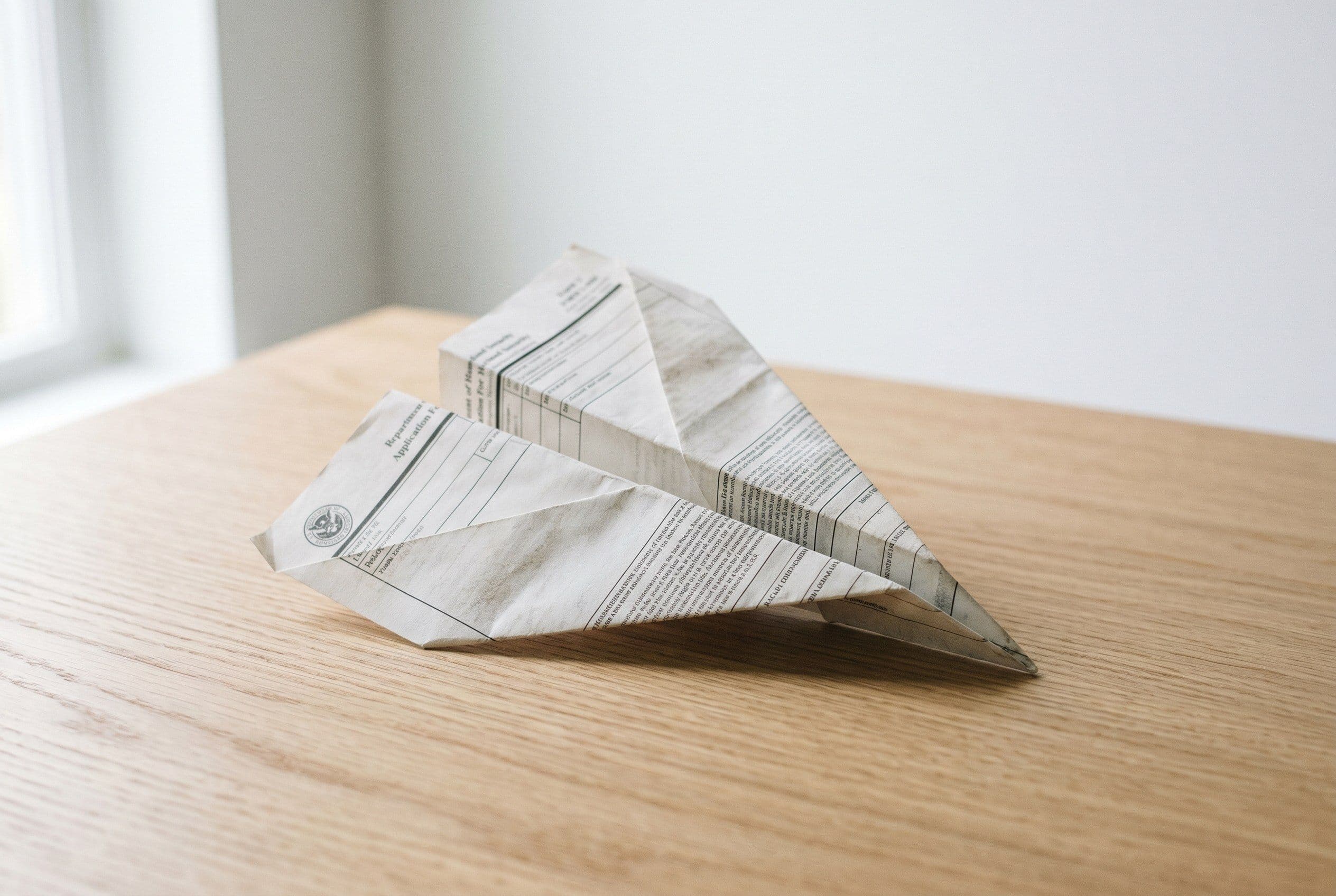 An I-130 immigration petition folded into a paper airplane on a wooden desk — USCIS policy memo PM-602-0199 explained
